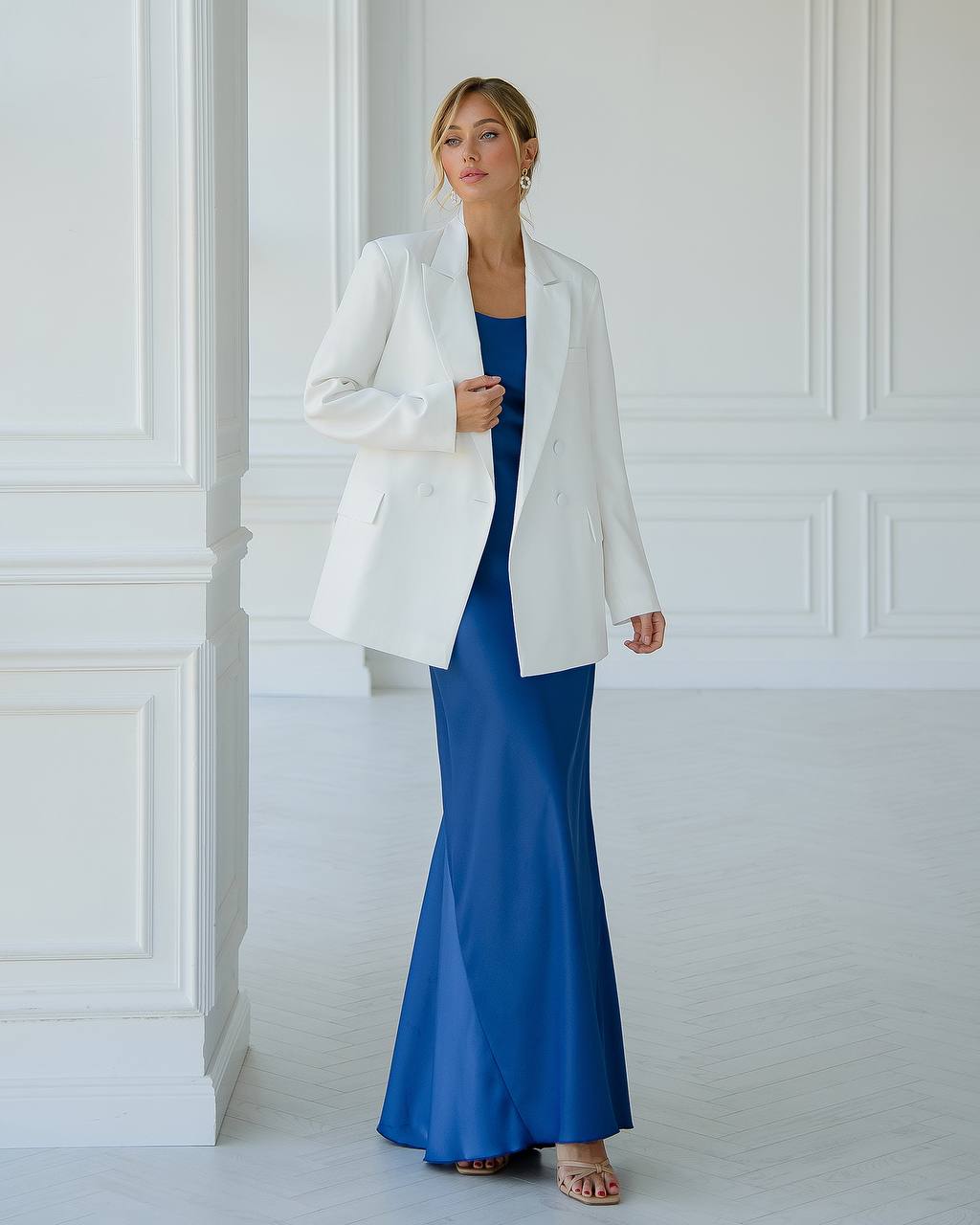 Woman wearing a white blazer over a blue dress against a white paneled wall.