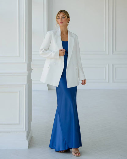 Woman wearing a white blazer over a blue dress against a white paneled wall.