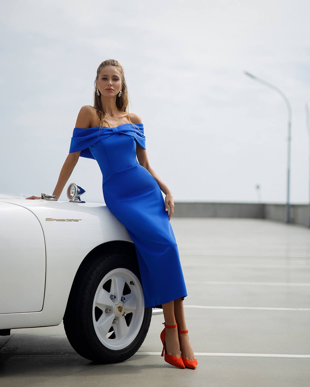 Woman in a blue off-shoulder dress sitting on a white car with a blurred background