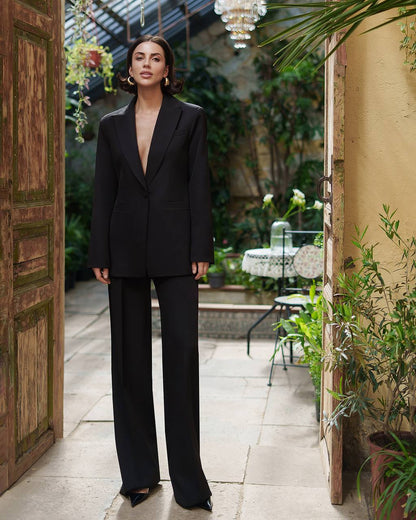 Woman in a black suit standing in an outdoor setting with plants and a wooden door.