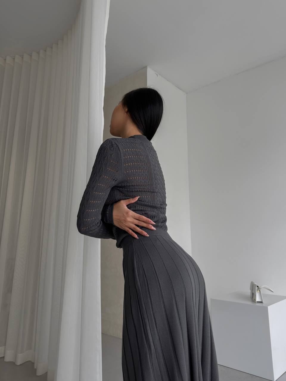 Woman in a long, dark gray dress standing in a minimalistic room with white walls and a white curtain.