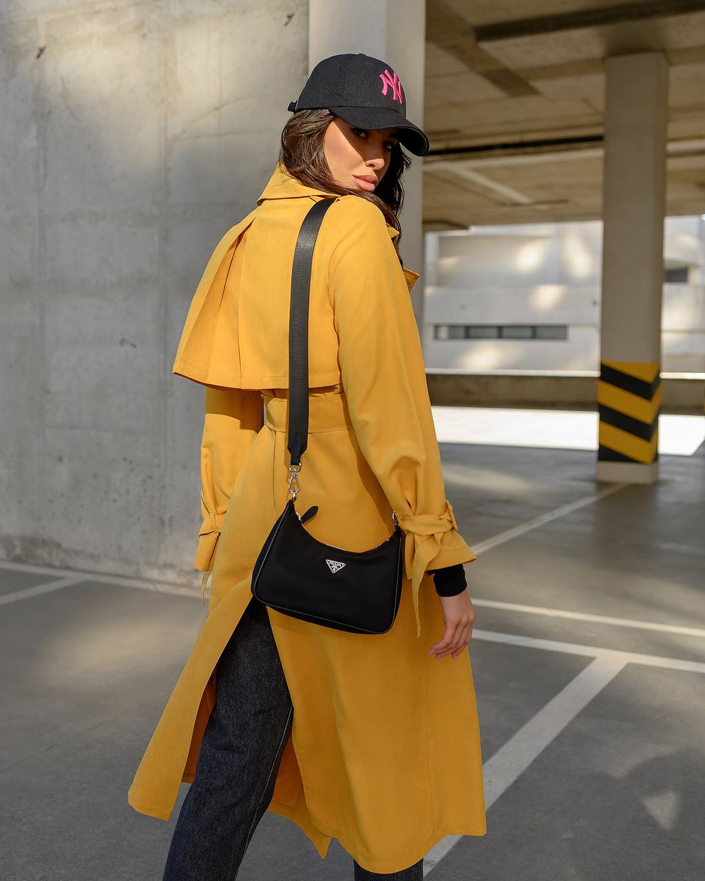 Person wearing a yellow coat and black cap, holding a black handbag in an urban setting.