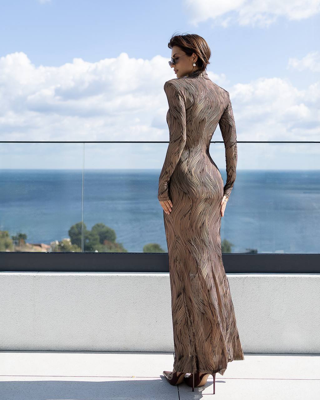 Woman in a patterned dress standing on a balcony with a scenic view of water and sky.