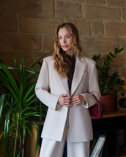 Woman in a beige suit standing in a room with plants and a brick wall.
