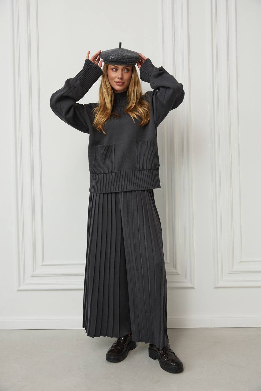 Woman wearing a black outfit with a pleated skirt against a white paneled wall.