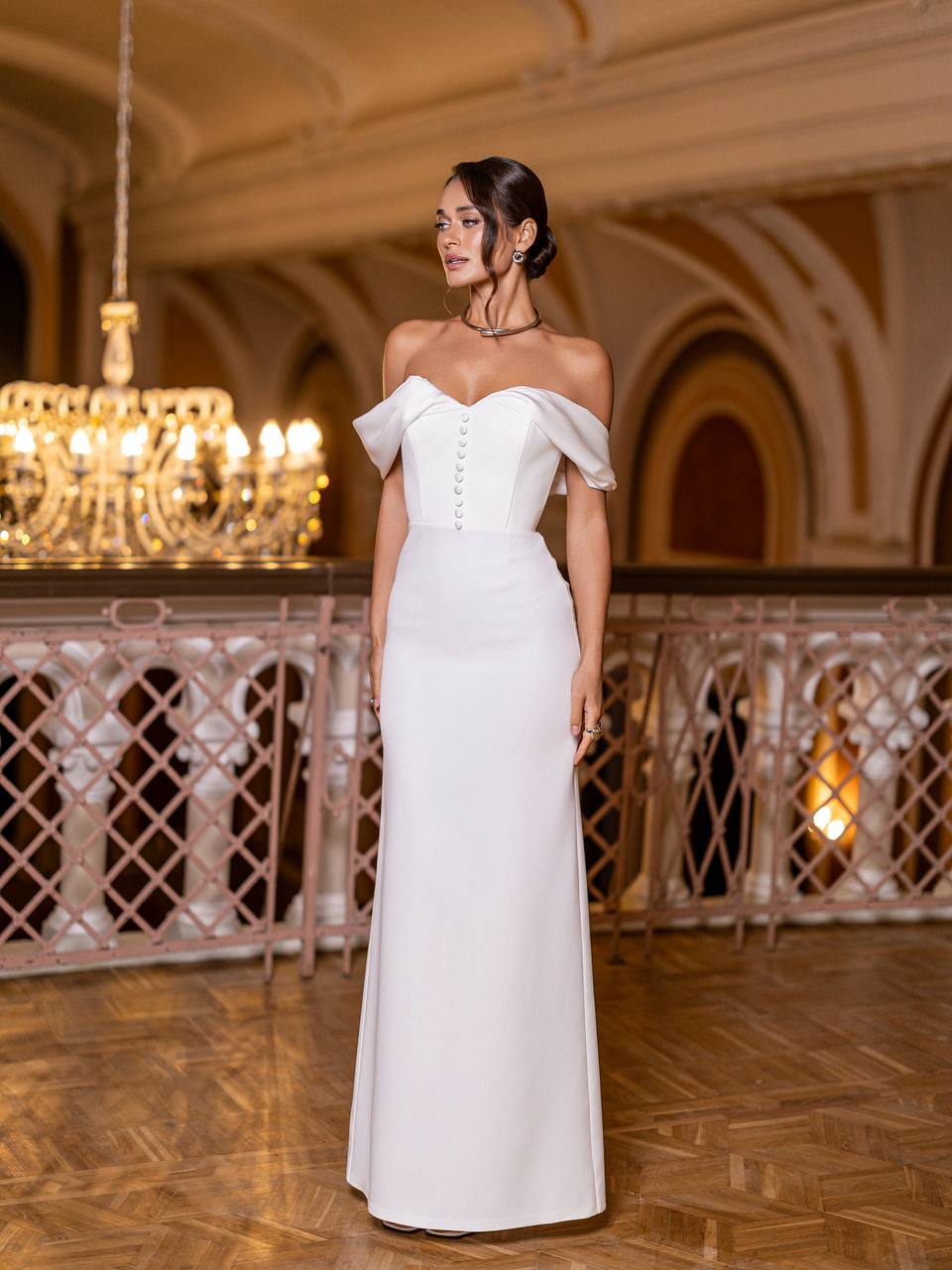 Woman in a white off-shoulder gown standing in an elegant indoor setting with chandeliers.