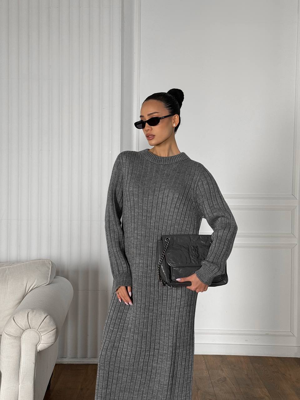 Woman in a gray ribbed dress and sweater holding a black clutch in a room with white walls and a white chair.
