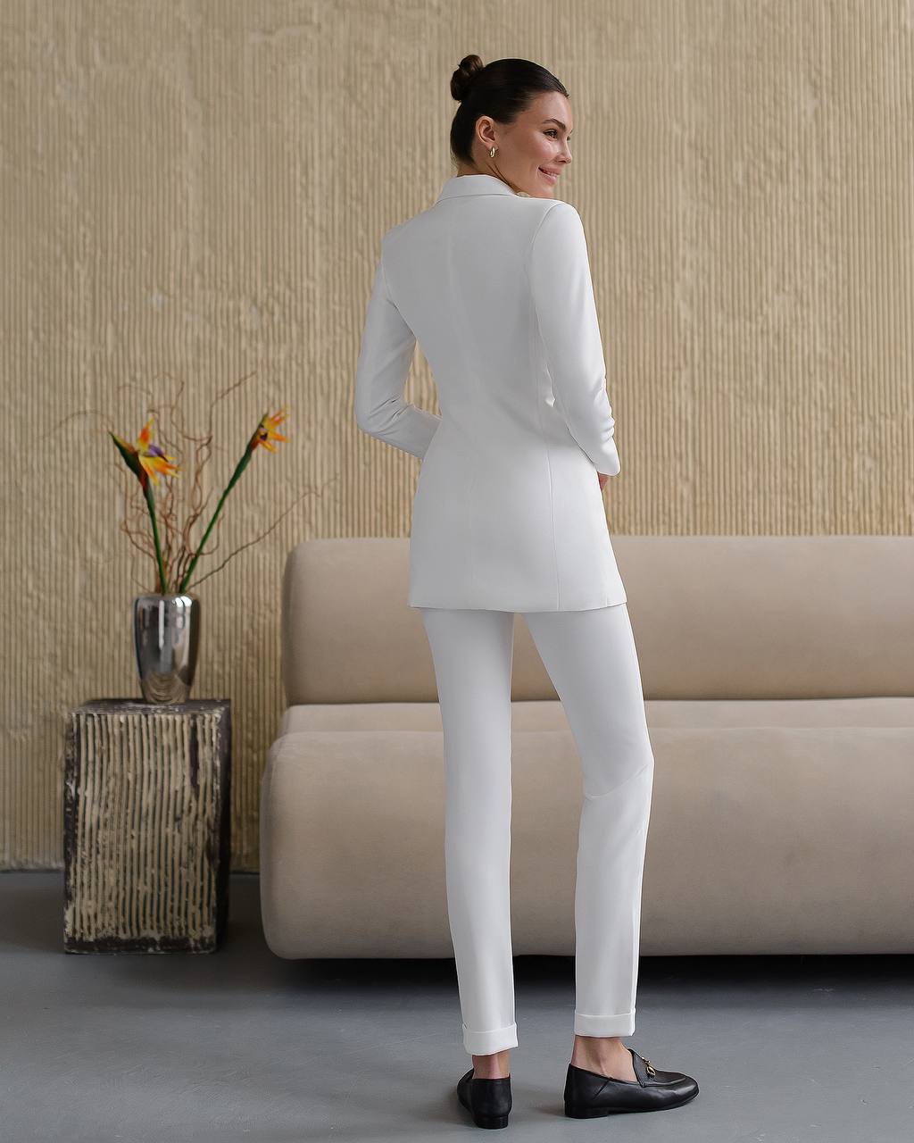 Woman in a white suit standing in a living room with a beige sofa and decorative elements.