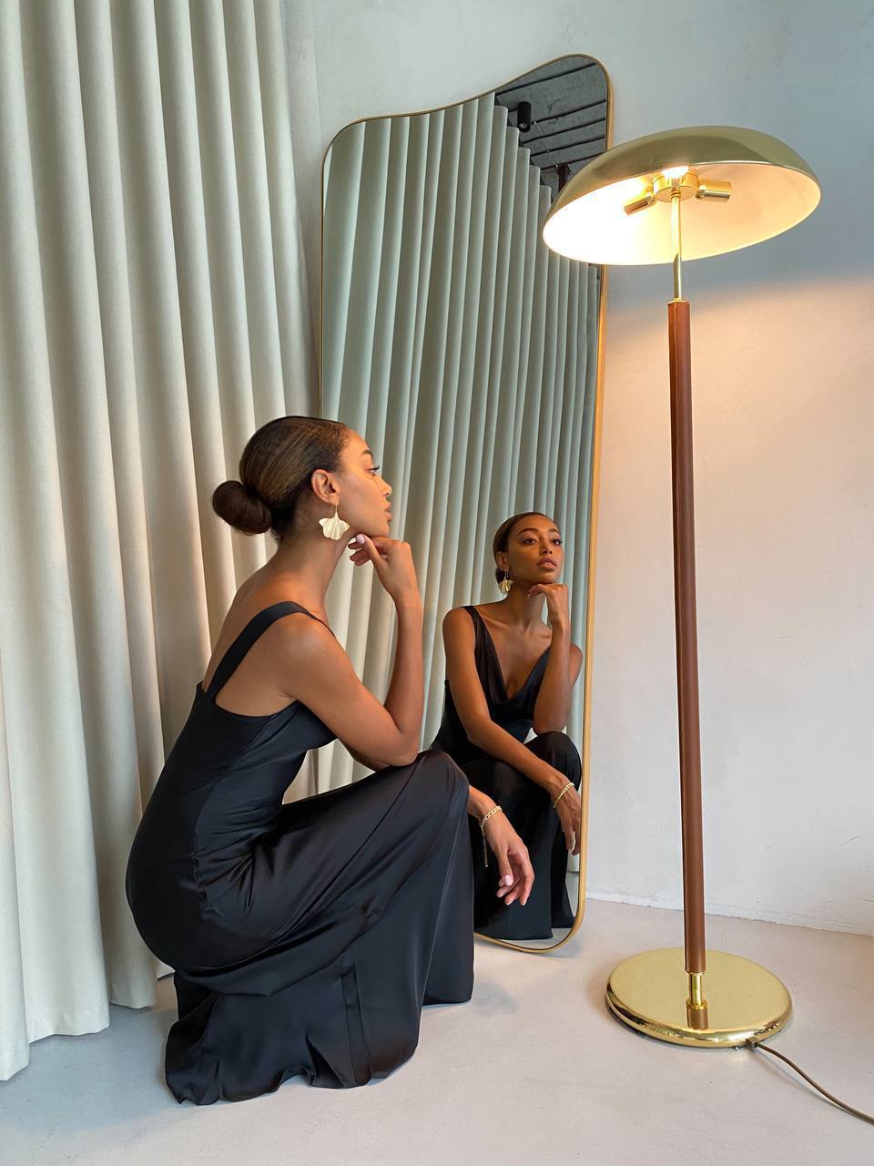 Woman in a black dress sitting on the floor next to a gold floor lamp and mirror.