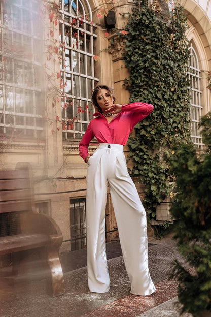 Woman in a red blouse and white pants standing in an outdoor setting with greenery and architectural elements.