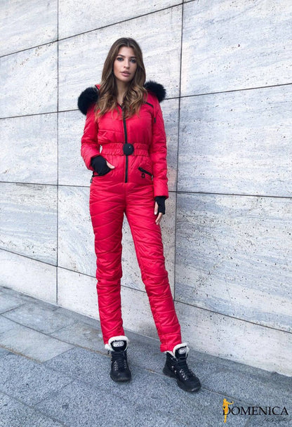 Woman wearing a red and black snowsuit standing against a gray wall.