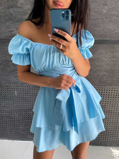 Woman wearing a light blue off-shoulder dress against a gray wall.