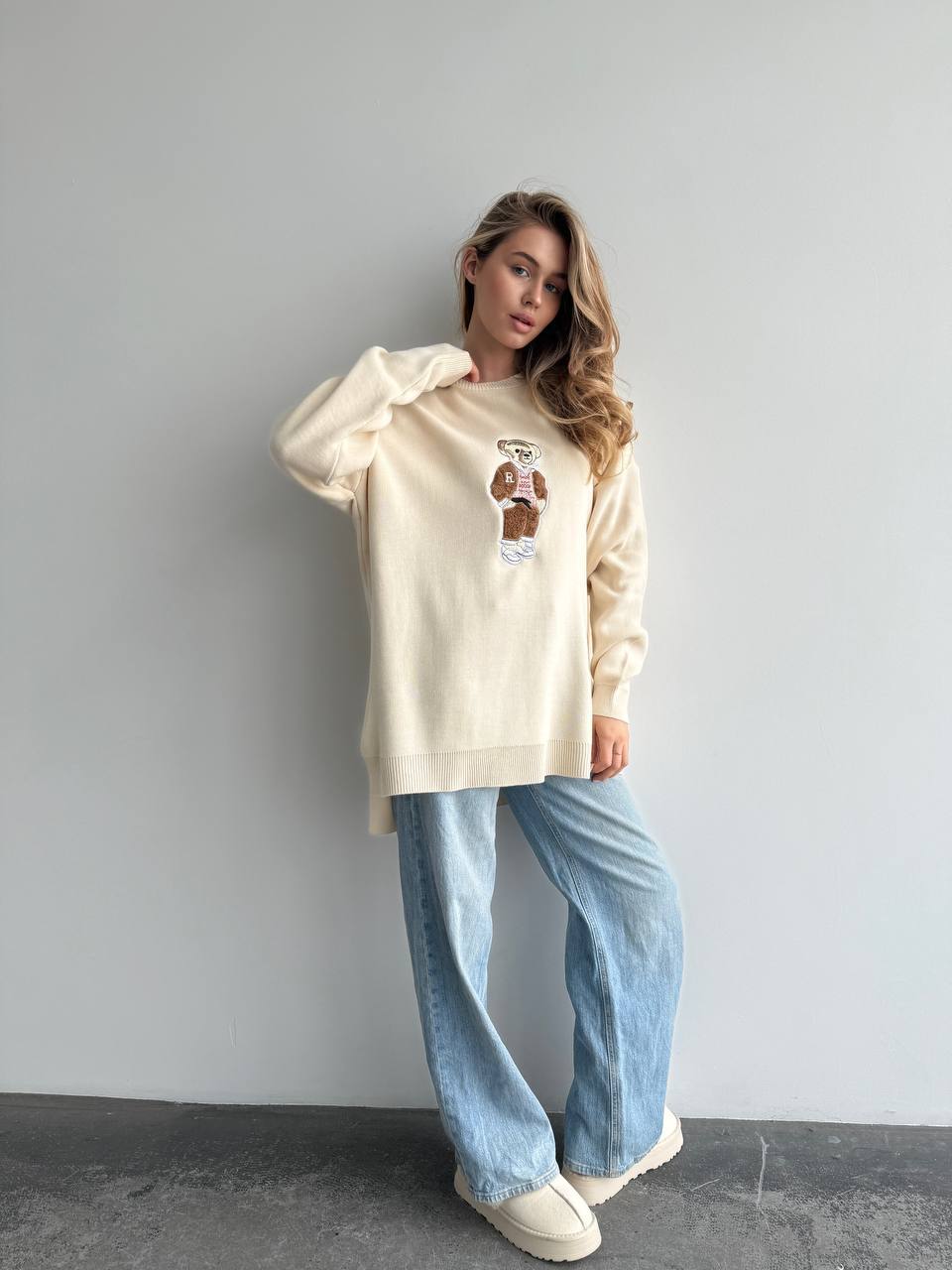 Woman wearing a cream sweatshirt with a graphic design and light blue jeans against a gray background