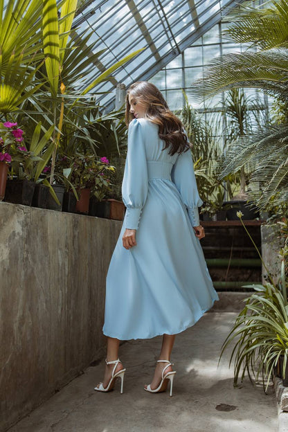Woman in a light blue dress standing in a greenhouse with plants and flowers around.