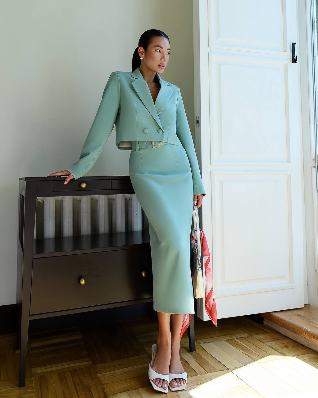 Woman in a light blue suit standing in a room with wooden flooring and white walls.