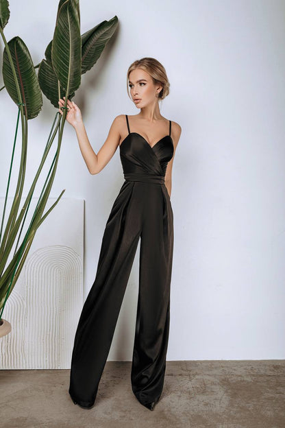 Woman wearing a black satin jumpsuit standing next to a plant against a white wall.