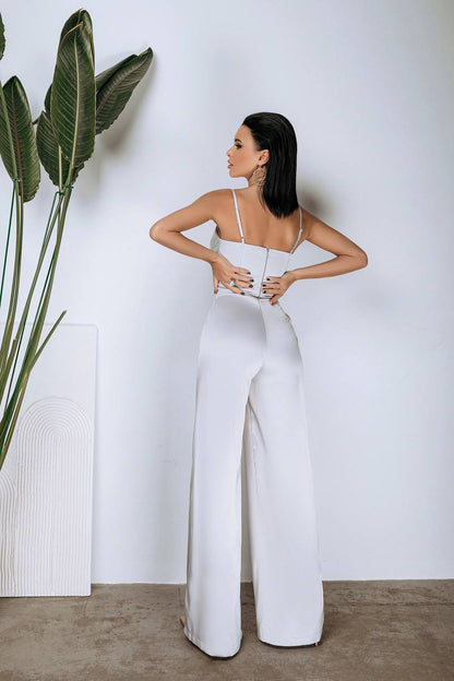 Woman wearing a white jumpsuit standing next to a plant against a plain wall.