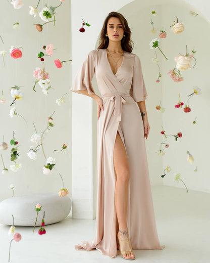 Woman in a long, light pink dress with a slit standing against a floral wall.