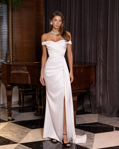 Woman in a white off-shoulder gown standing in an elegant room with a piano and dark curtains.
