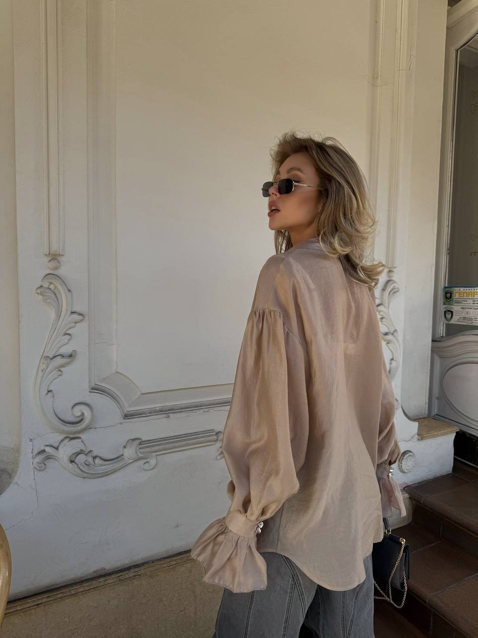 Woman wearing a beige blouse and sunglasses in an indoor setting with decorative wall.