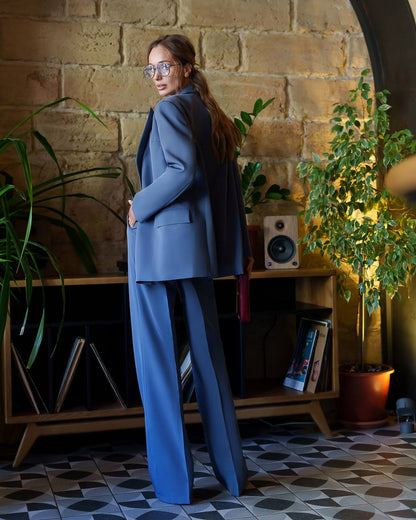 Woman in a blue suit standing in a room with plants and a wooden shelf.