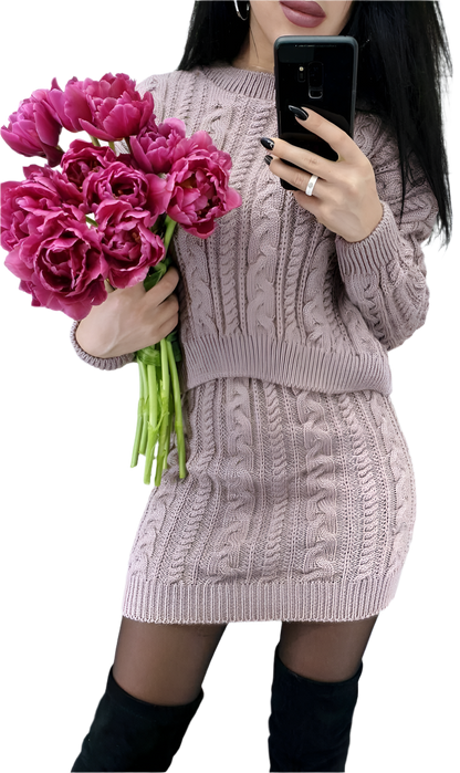 Person wearing a cable knit sweater holding pink flowers and a phone.