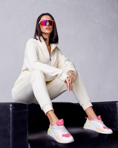 Person wearing a white tracksuit and colorful sneakers sitting on a black chair against a gray background.