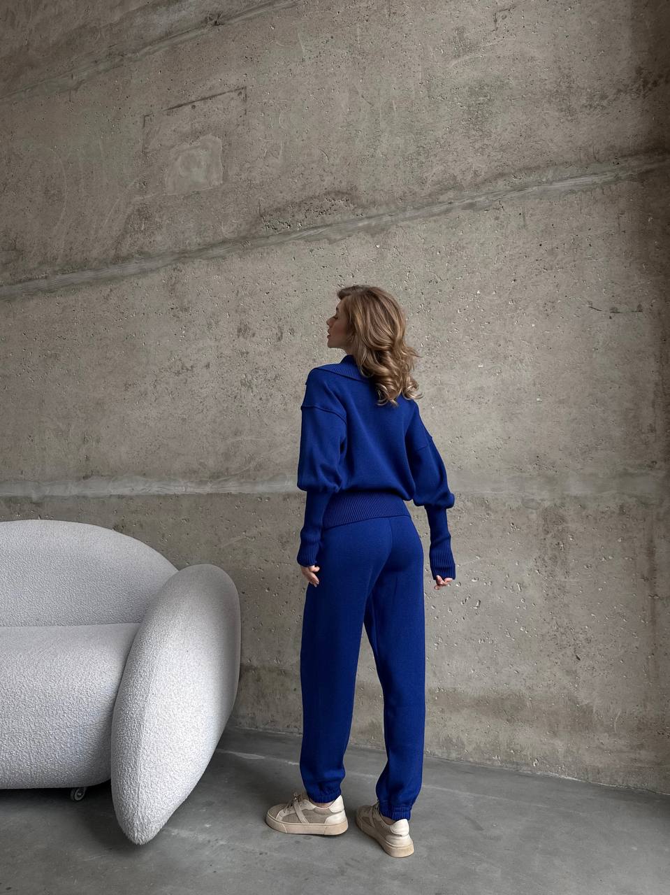 Person in a blue outfit standing in a room with a modern white chair against a concrete wall.