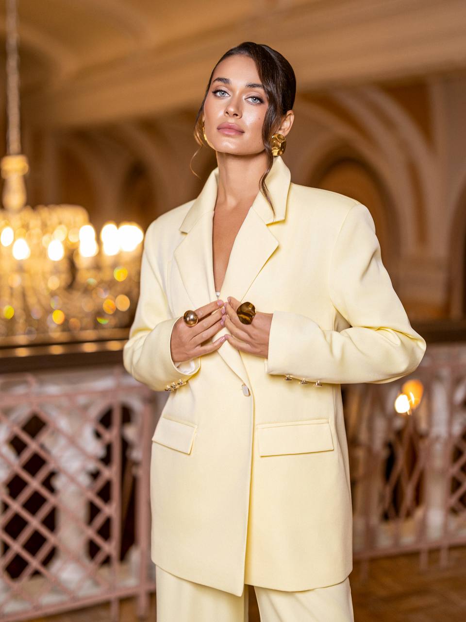 Woman in a light yellow suit standing in an elegant interior setting with chandeliers.