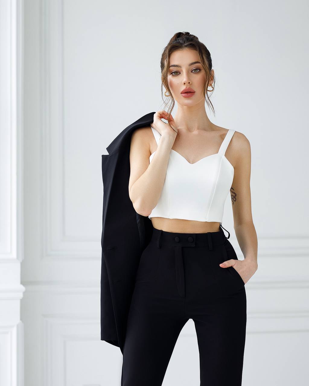 Woman wearing a white top and black pants with a black jacket over her shoulder against a white background