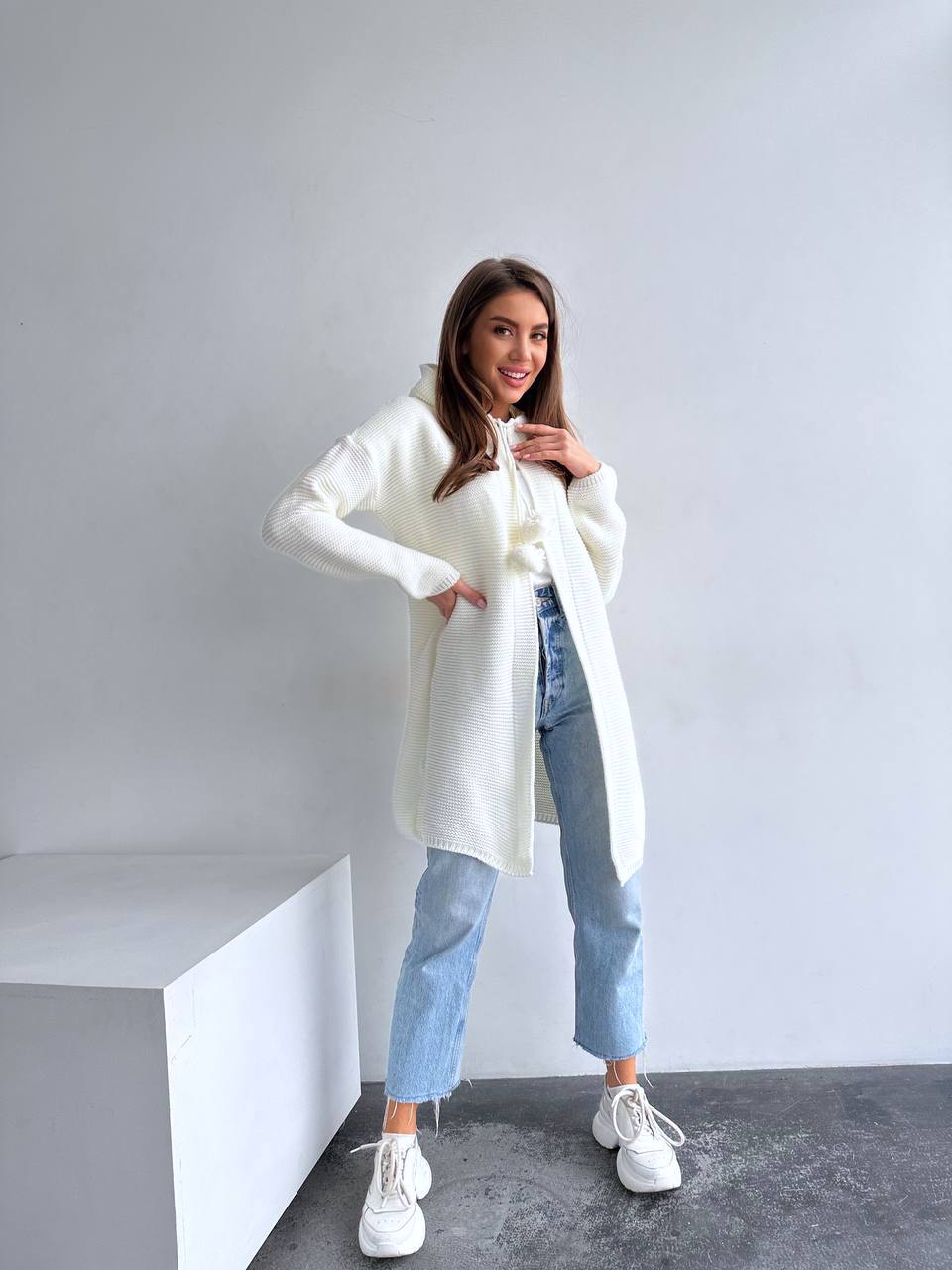 Woman wearing a white cardigan, blue jeans, and white sneakers in a minimalistic setting.