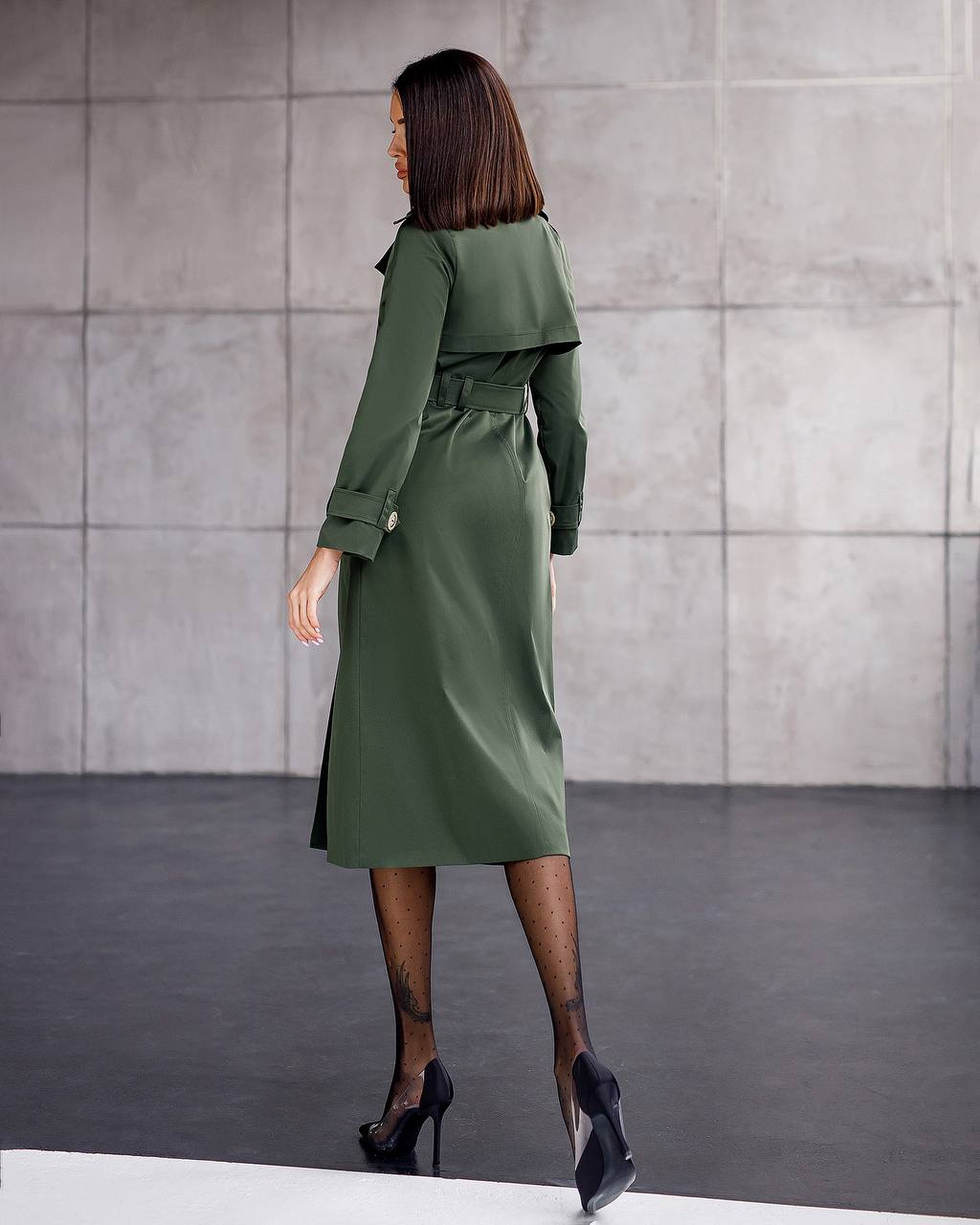 Woman wearing a green trench coat standing against a gray tiled wall.