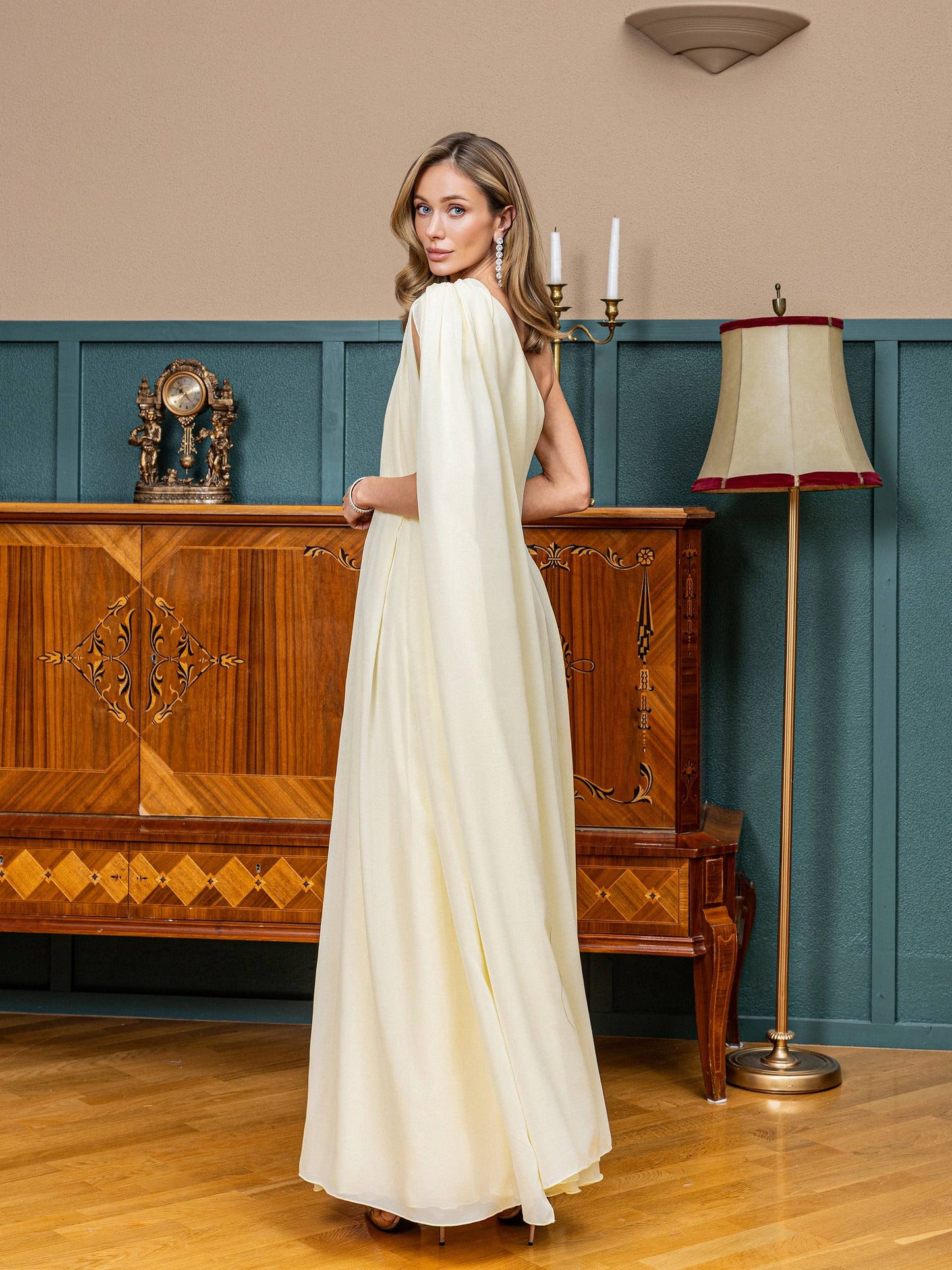 Woman in a white dress standing in a room with wooden furniture and a lamp.