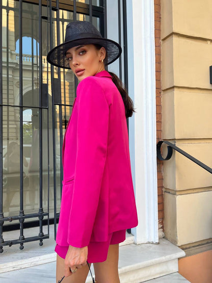 Woman in a bright pink outfit and black hat standing outside a building.