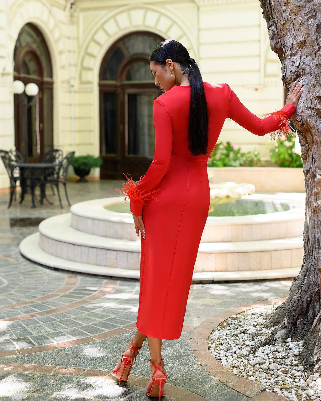 Woman in a red dress standing in an outdoor setting with a fountain and building.