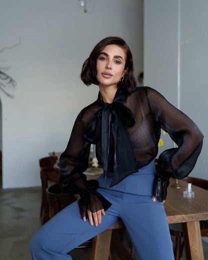 Woman wearing a black sheer blouse with a bow and blue pants in an indoor setting.