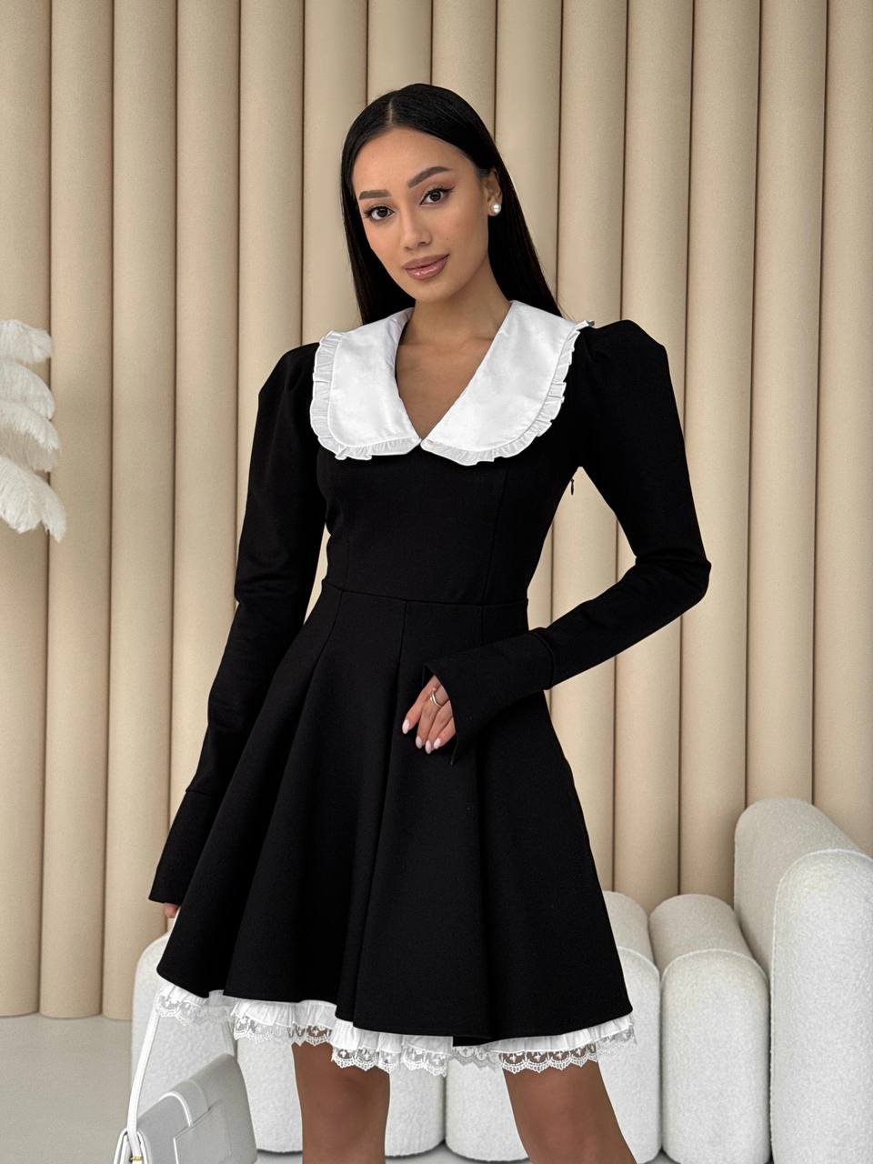 Woman wearing a black dress with white collar and lace trim in a room with beige curtains.