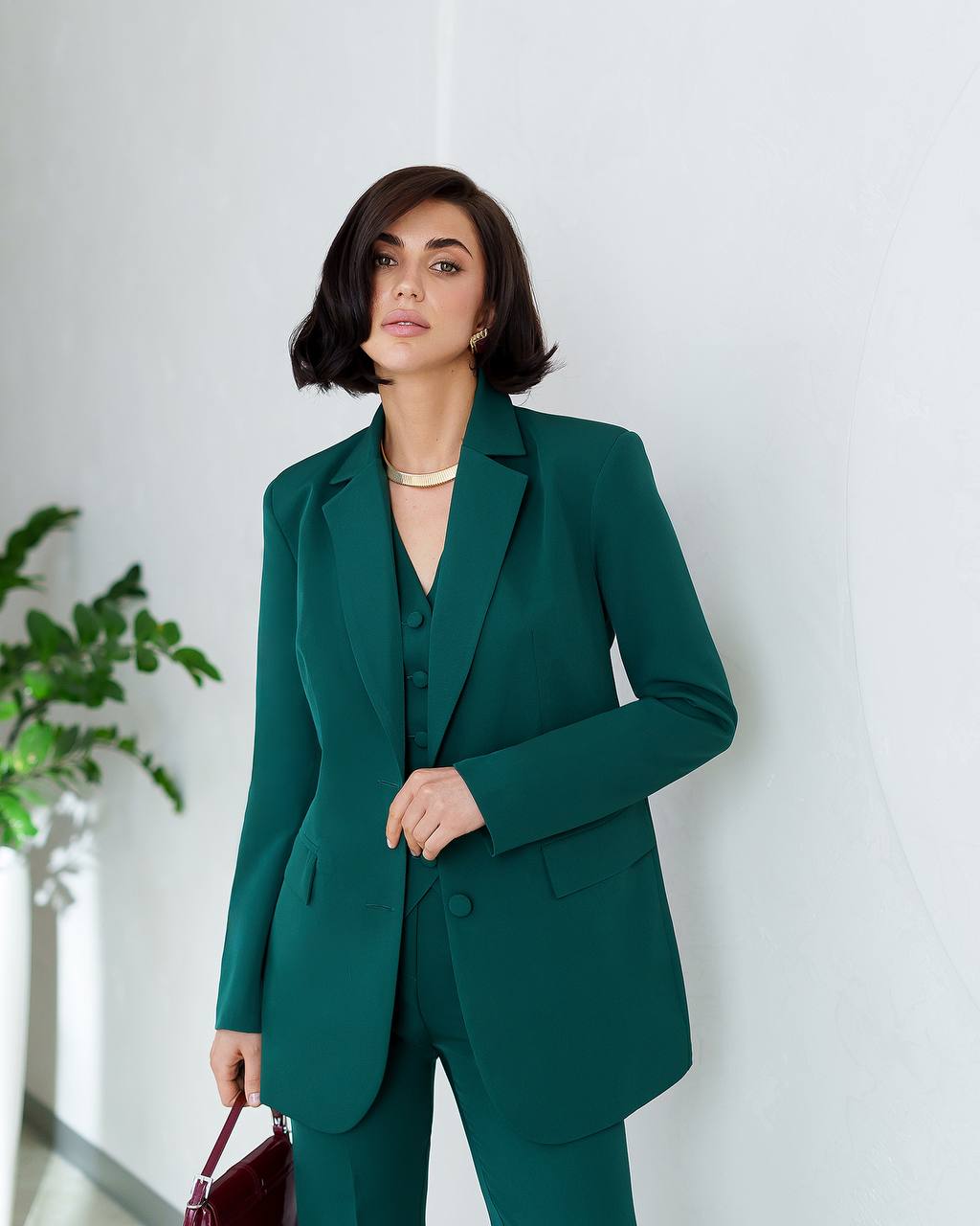 Woman wearing a green suit standing against a white wall with a plant to the side.