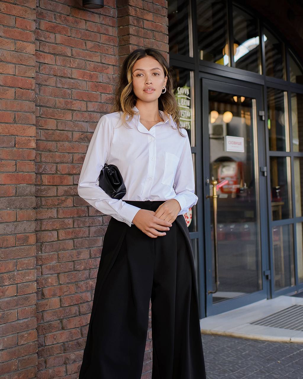 Woman in a white shirt and black pants standing against a brick wall.