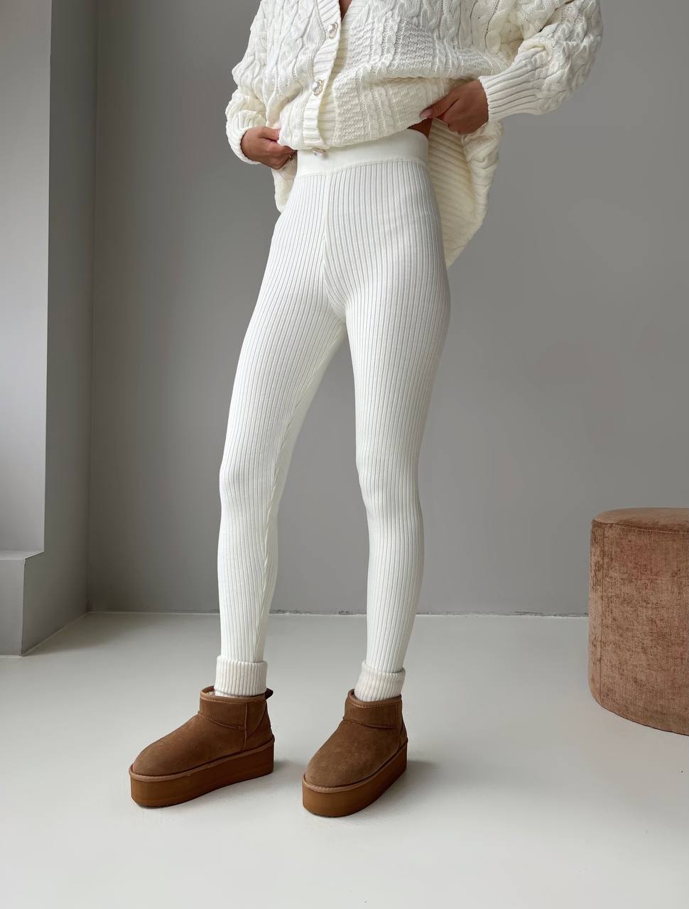 Person wearing a white knit sweater, leggings, and brown boots in a minimalistic setting.