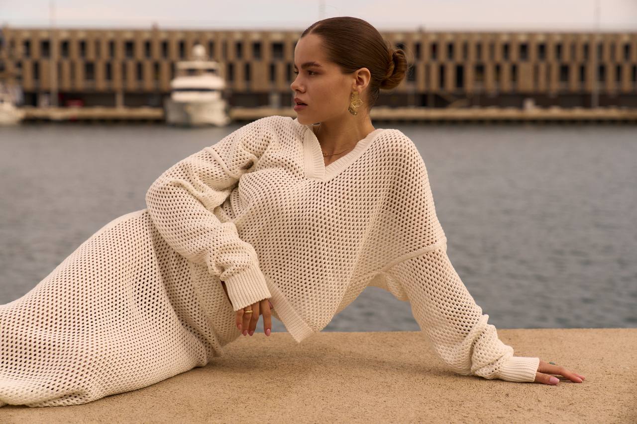 Woman in a beige knit dress sitting by a waterfront with a building in the background