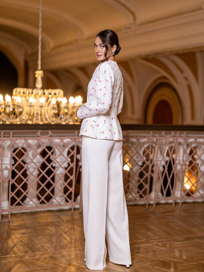 Woman in a floral blouse and white pants standing in an elegant interior setting with chandeliers.