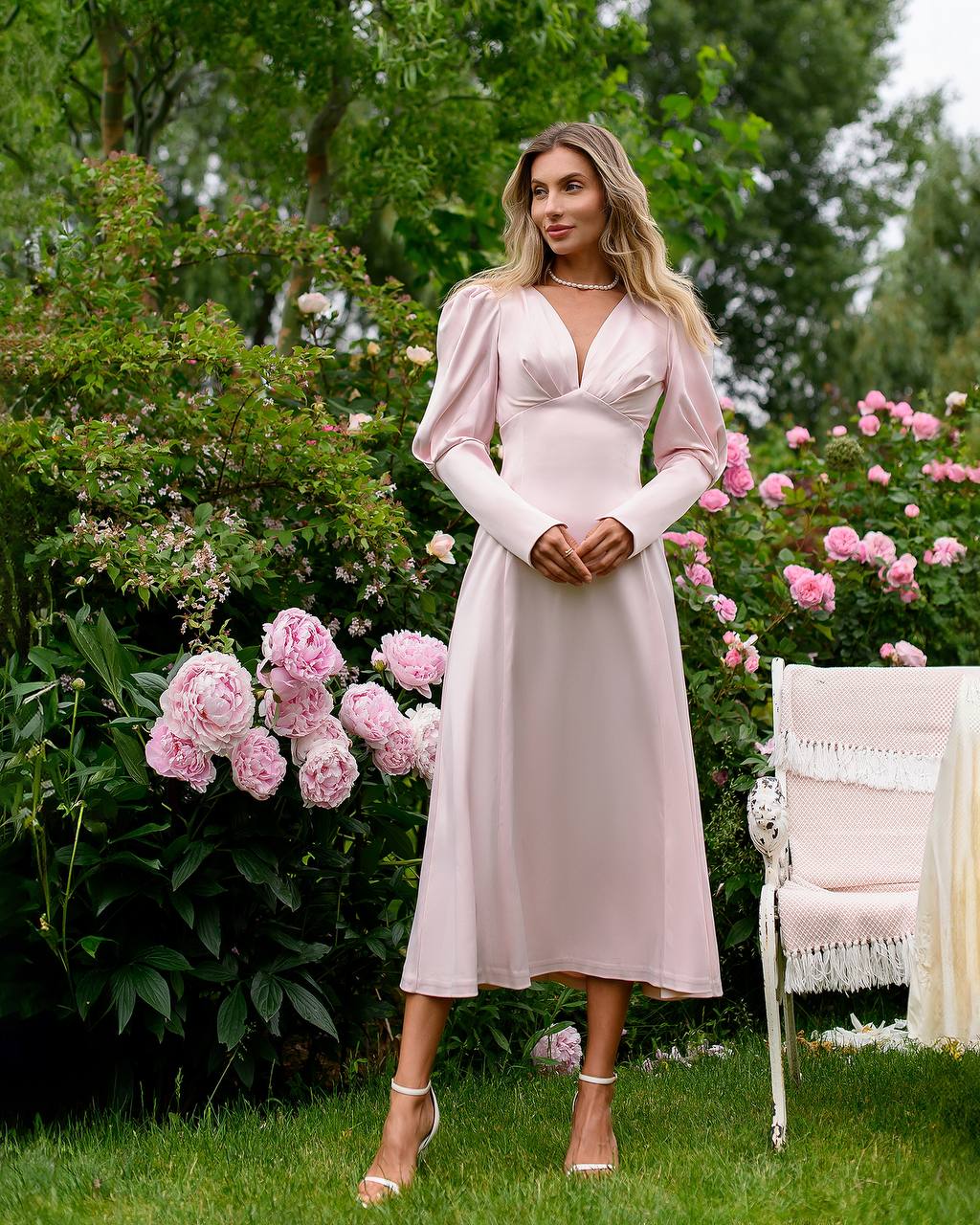 Woman in a pink dress standing in a garden with pink flowers and a white chair.