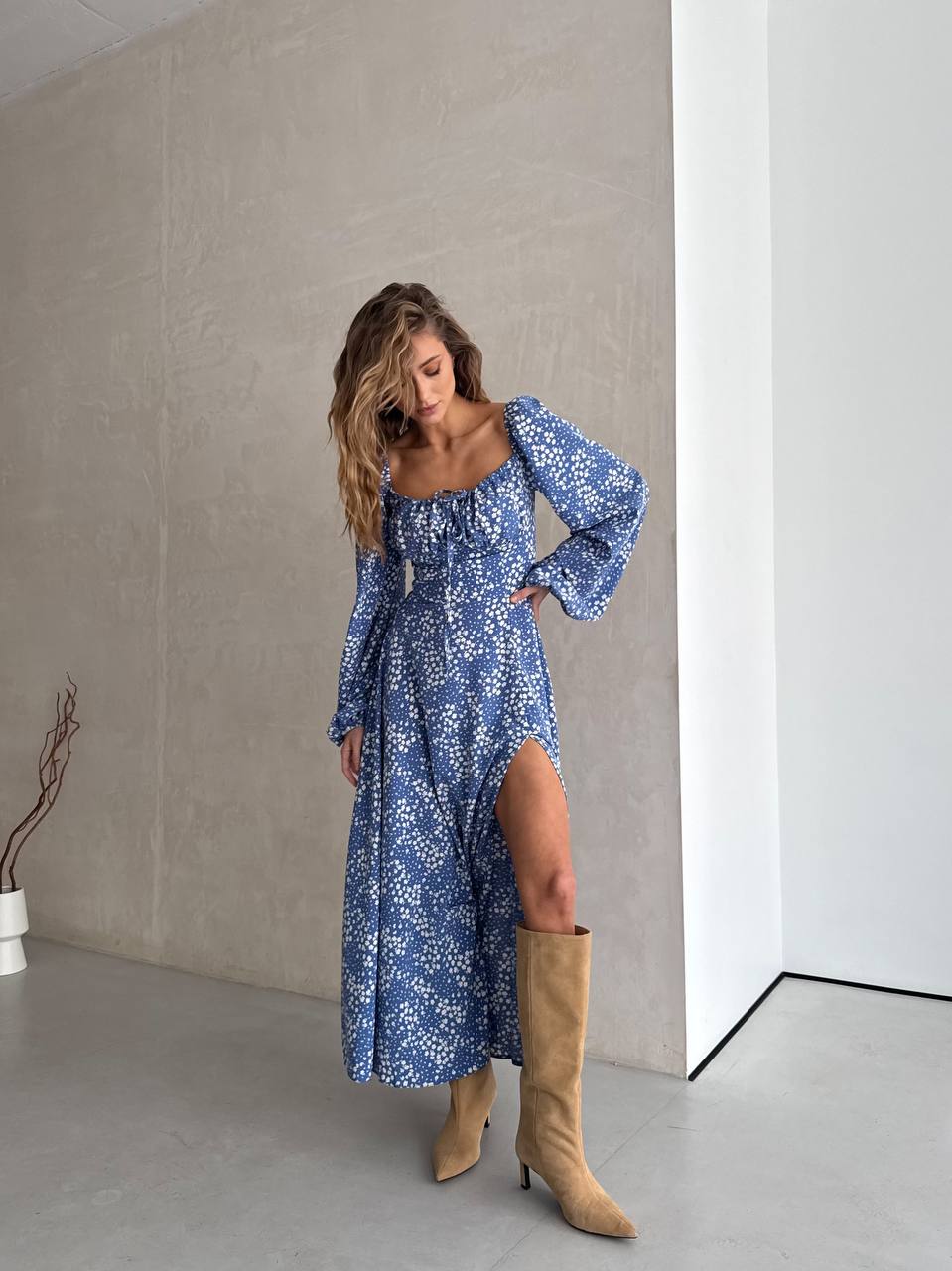 Woman wearing a blue floral dress with long sleeves and a slit, standing against a neutral wall.