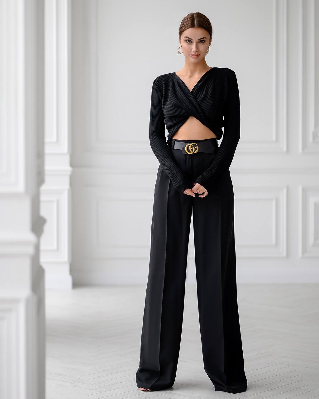 Woman wearing a black outfit with a Gucci belt against a white paneled wall.