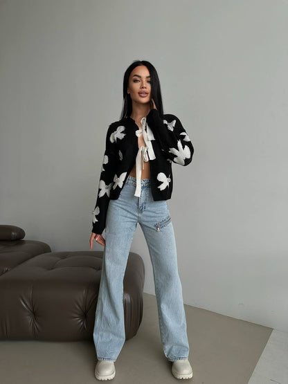 Woman wearing a black cardigan with white floral patterns, white top, and blue jeans indoors.