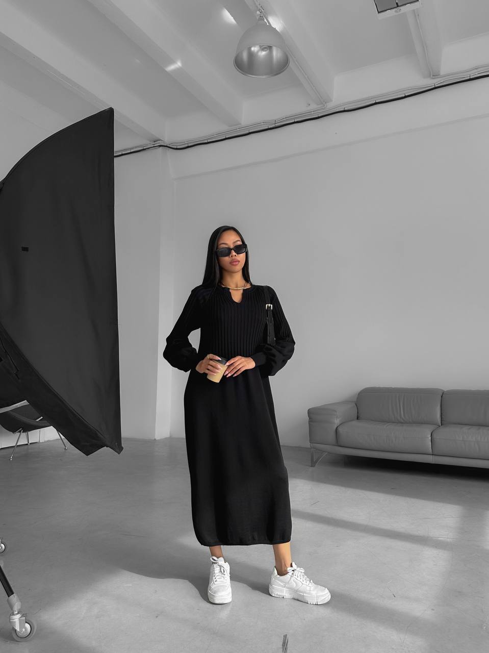 Person wearing a black dress and sunglasses in a studio setting with gray furniture.