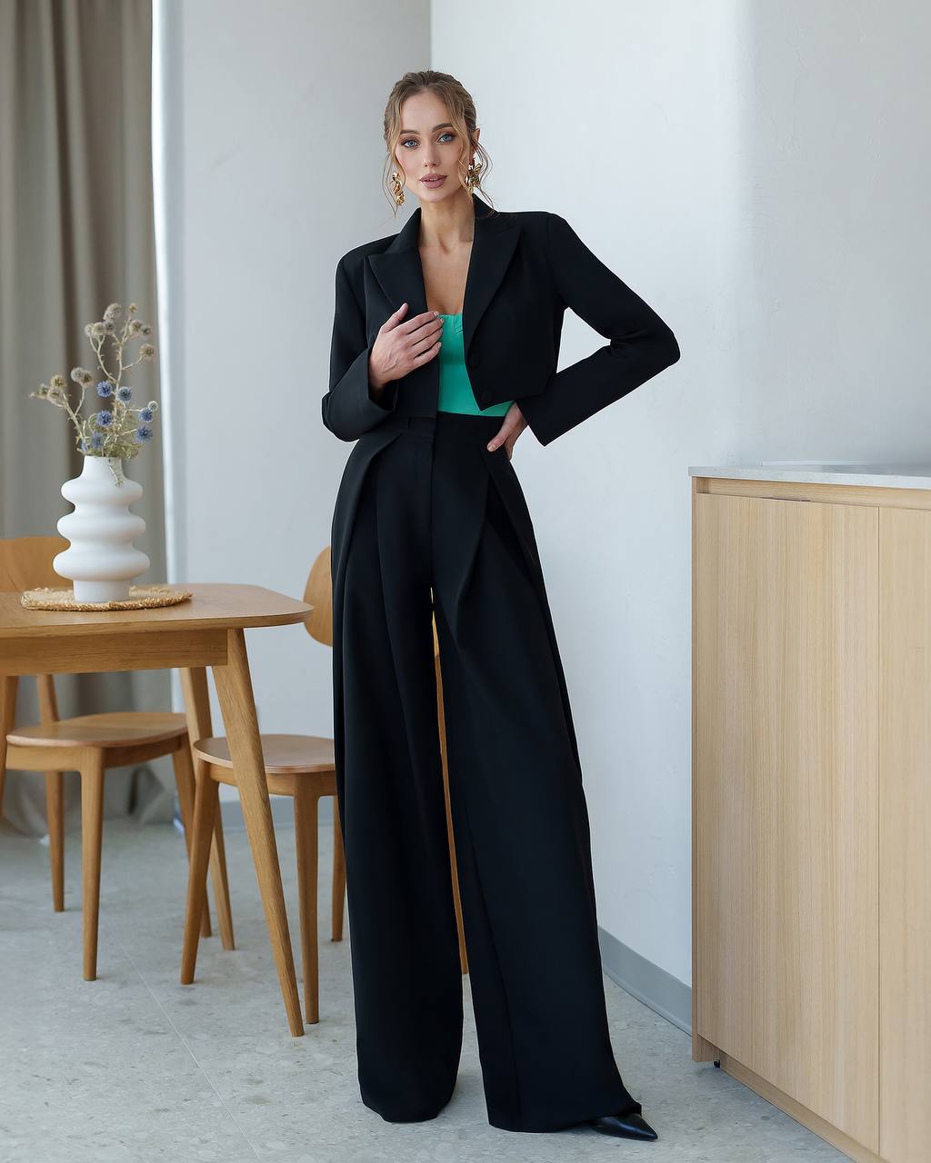 Woman in a black coat and green top standing in a modern room with wooden furniture.