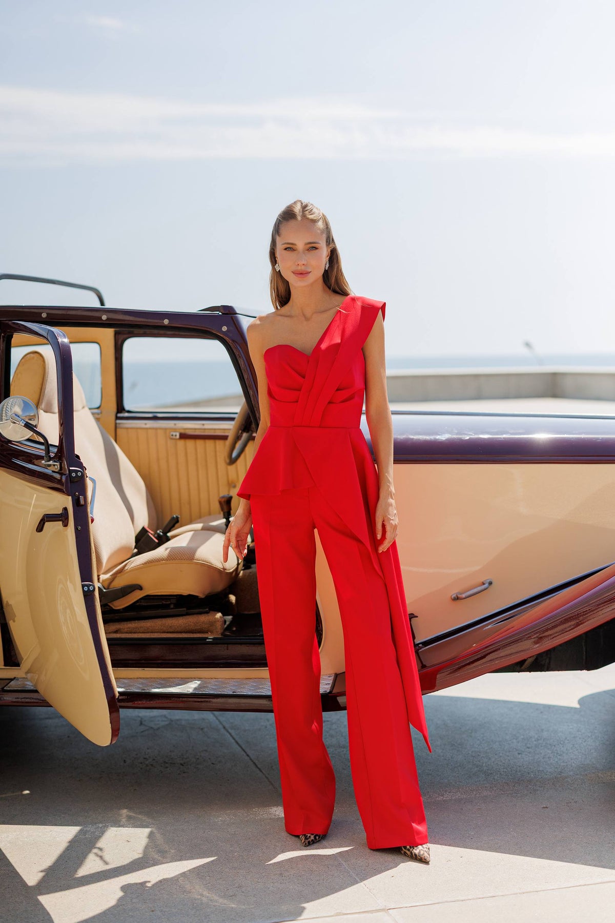 A woman stands outdoors beside an open vintage car wearing a bright red one-shoulder jumpsuit with wide legs and a peplum waist. The background shows a pale sky and a bit of water.