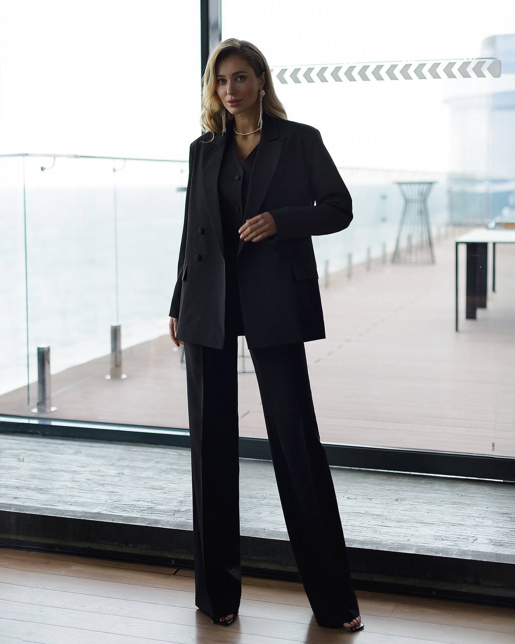 Woman in a black suit standing in a modern indoor setting with large windows.